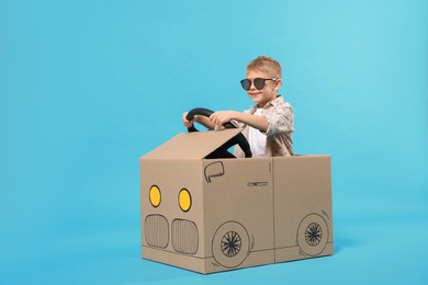 Little boy in sunglasses driving car made of cardboard on light blue background