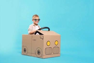 Photo of Little boy in sunglasses driving car made of cardboard on light blue background. Space for text