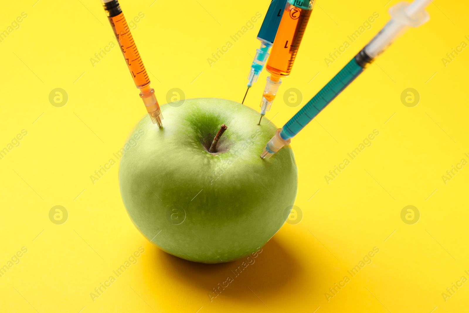 Photo of GMO concept. Green apple with different syringes on yellow background, closeup