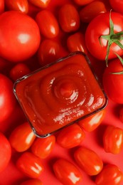 Photo of Ketchup in bowl and tomatoes on red background, top view