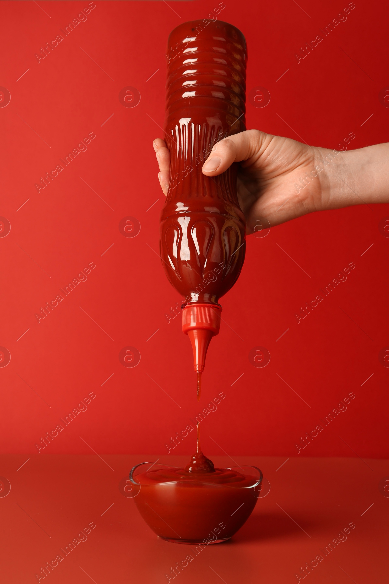 Photo of Woman squeezing ketchup from bottle into bowl on red background, closeup