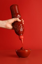 Photo of Woman squeezing ketchup from bottle into bowl on red background, closeup