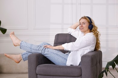 Smiling woman in stylish jeans listening to music on armchair at home