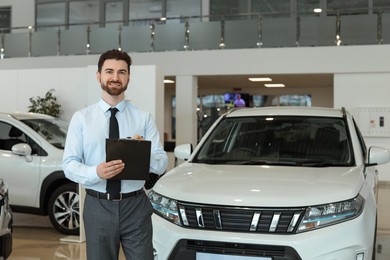 Photo of Happy salesman with clipboard near new cars in salon