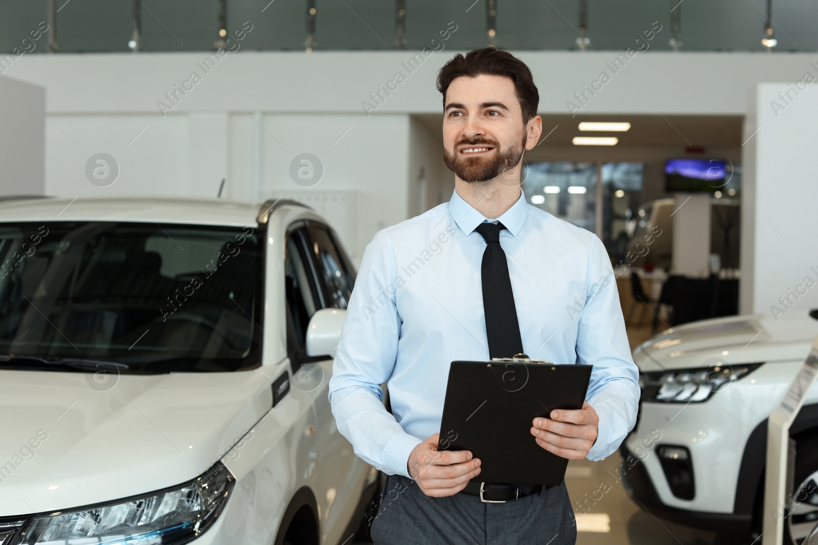 Photo of Happy salesman with clipboard near new cars in salon