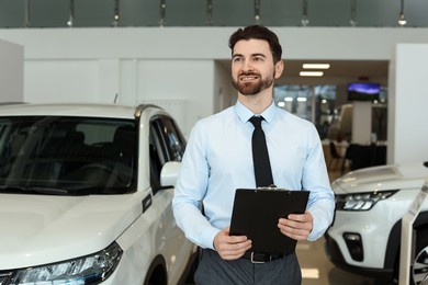 Photo of Happy salesman with clipboard near new cars in salon