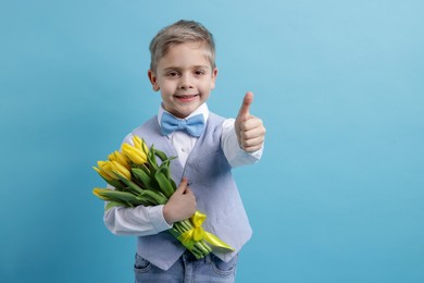 Photo of Cute little boy with bouquet of tulips showing thumbs up on light blue background