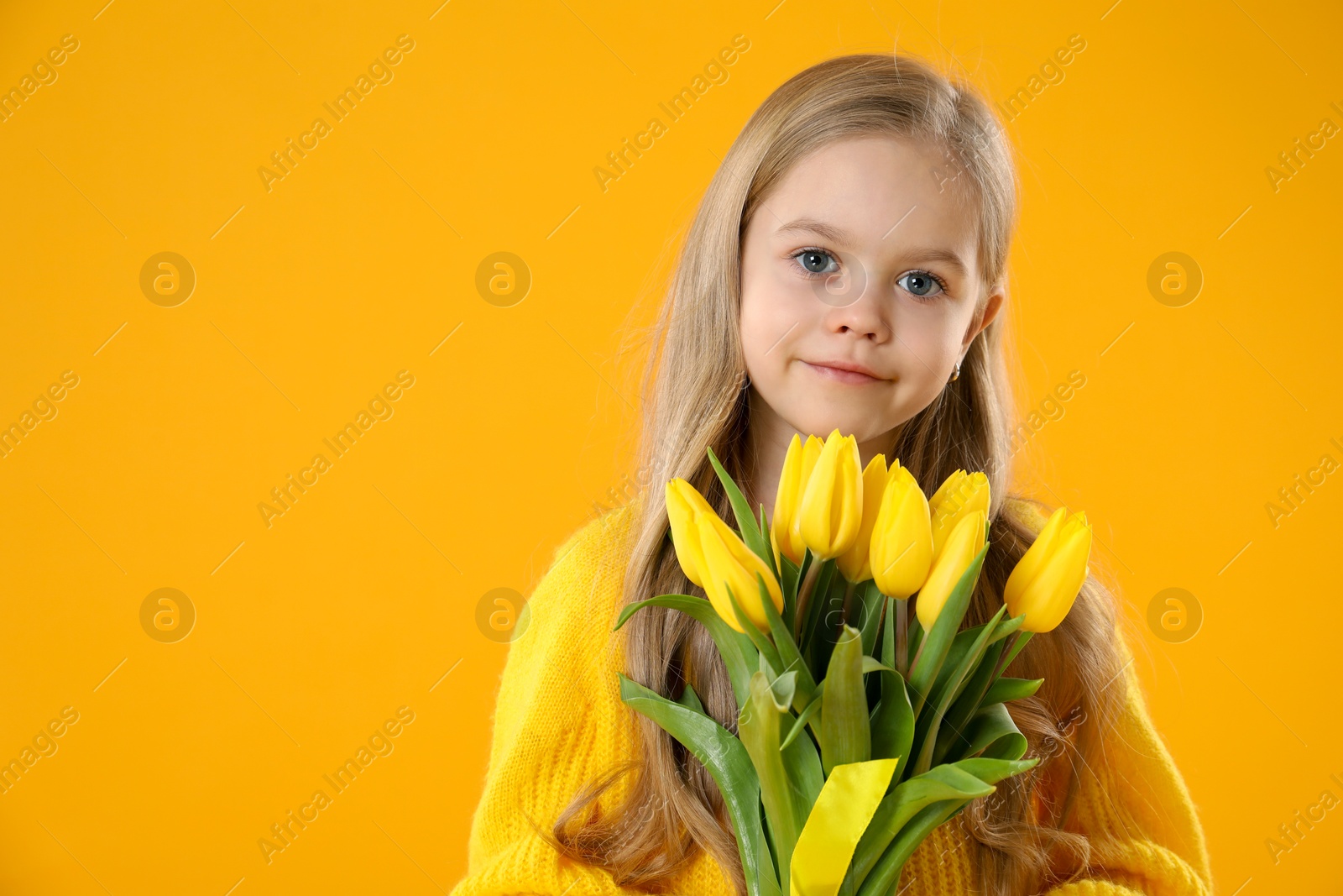 Photo of Cute little girl with beautiful yellow tulips on orange background, space for text. Spring season