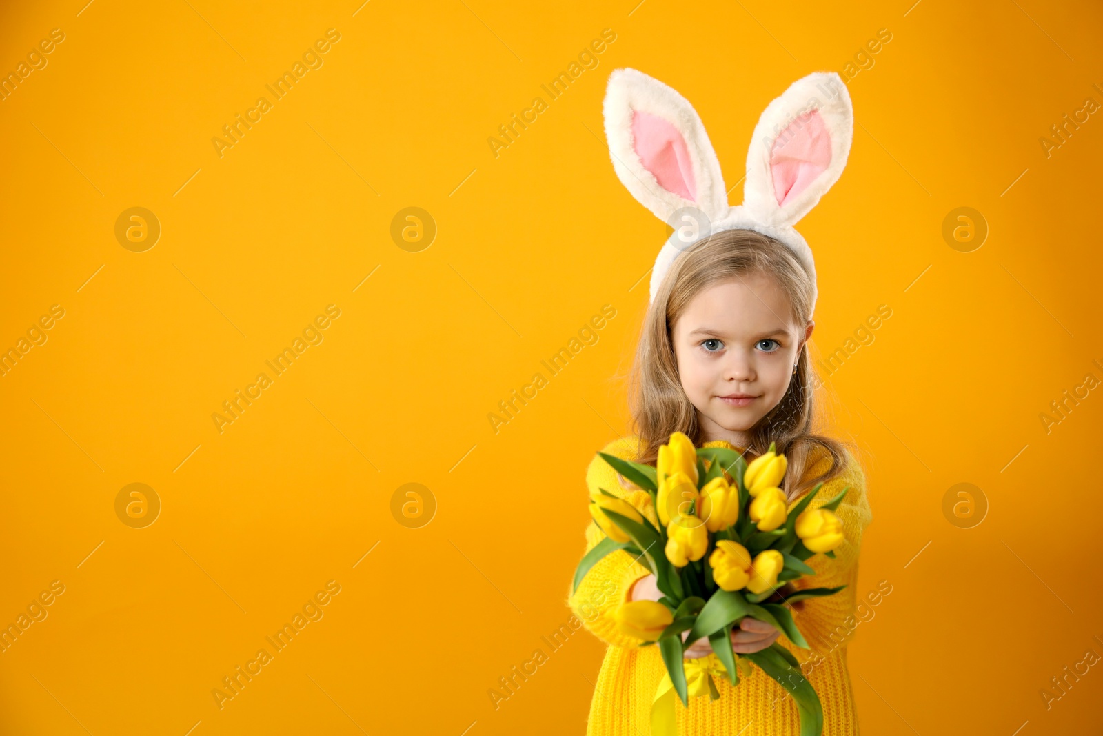 Photo of Cute little girl with bunny ears and tulips on orange background, space for text. Easter celebration