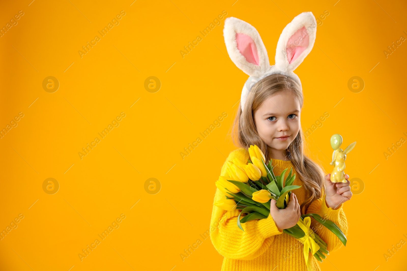 Photo of Cute little girl with bunny ears and tulips on orange background, space for text. Easter celebration