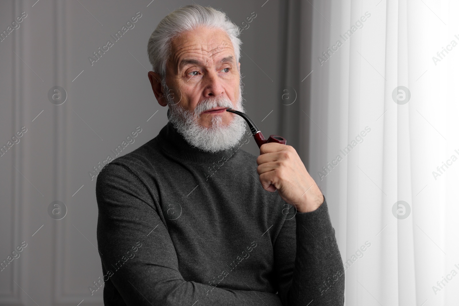 Photo of Portrait of handsome bearded man with tobacco pipe near window indoors