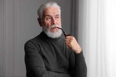 Photo of Portrait of handsome bearded man with tobacco pipe near window indoors