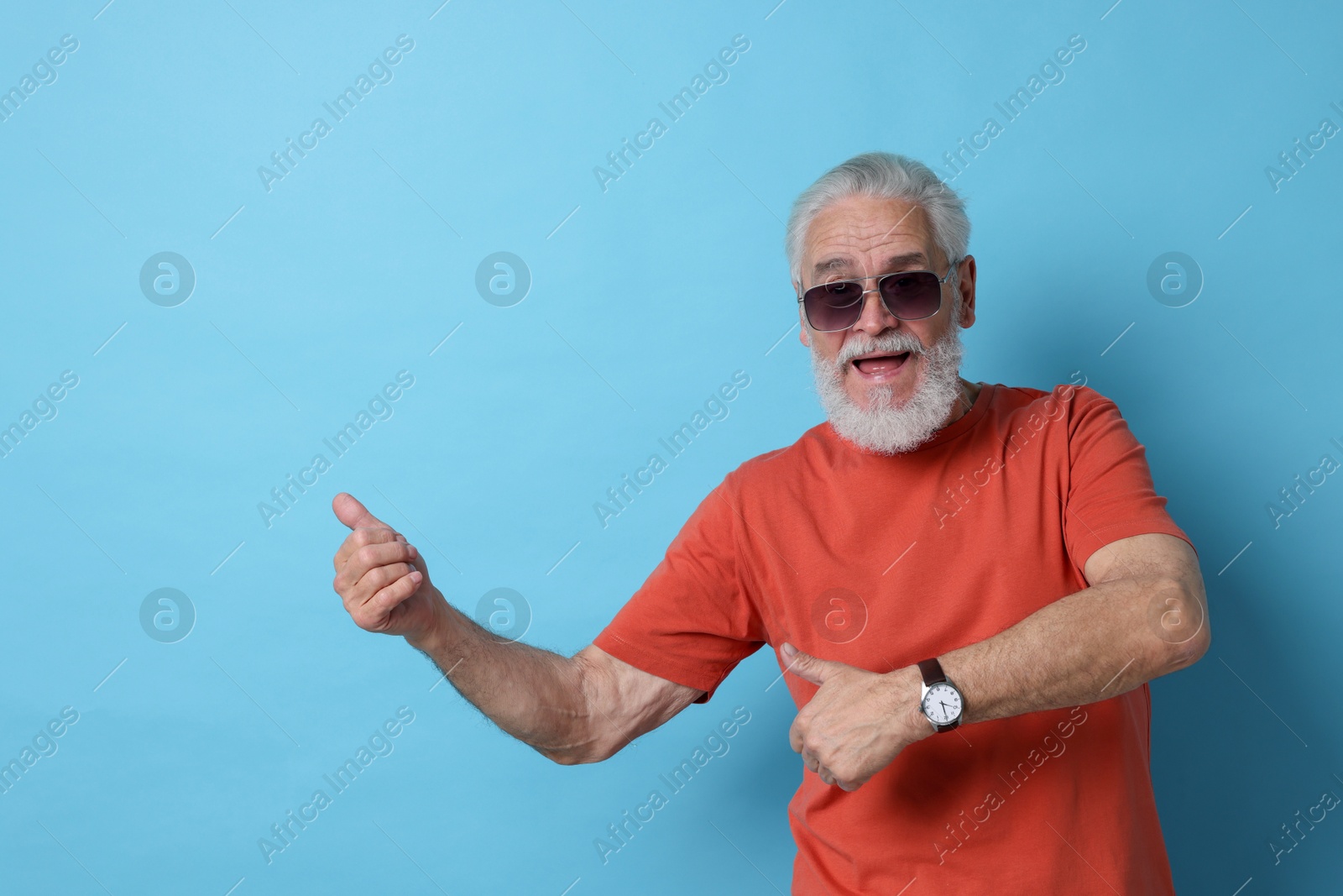 Photo of Emotional senior man in sunglasses pointing at something on light blue background. Space for text