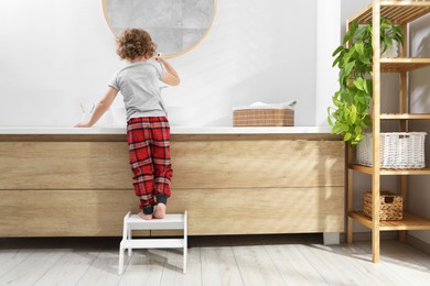 Photo of Little boy standing on step stool near bathroom vanity indoors