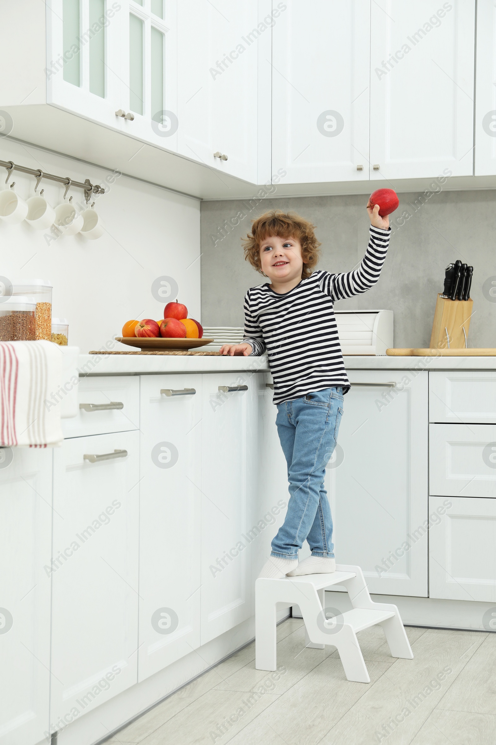 Photo of Little boy with apple standing on step stool in kitchen