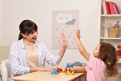 Photo of Autism therapy. Smiling psychologist giving high five to little girl at table in mental health center