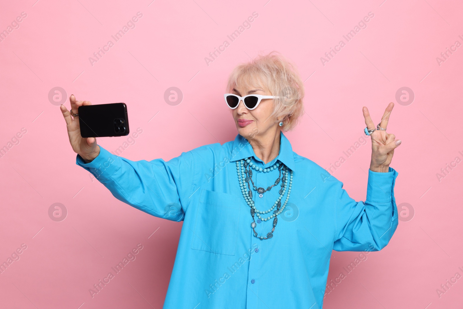 Photo of Cool grandmother showing V-sign while taking selfie on pink background