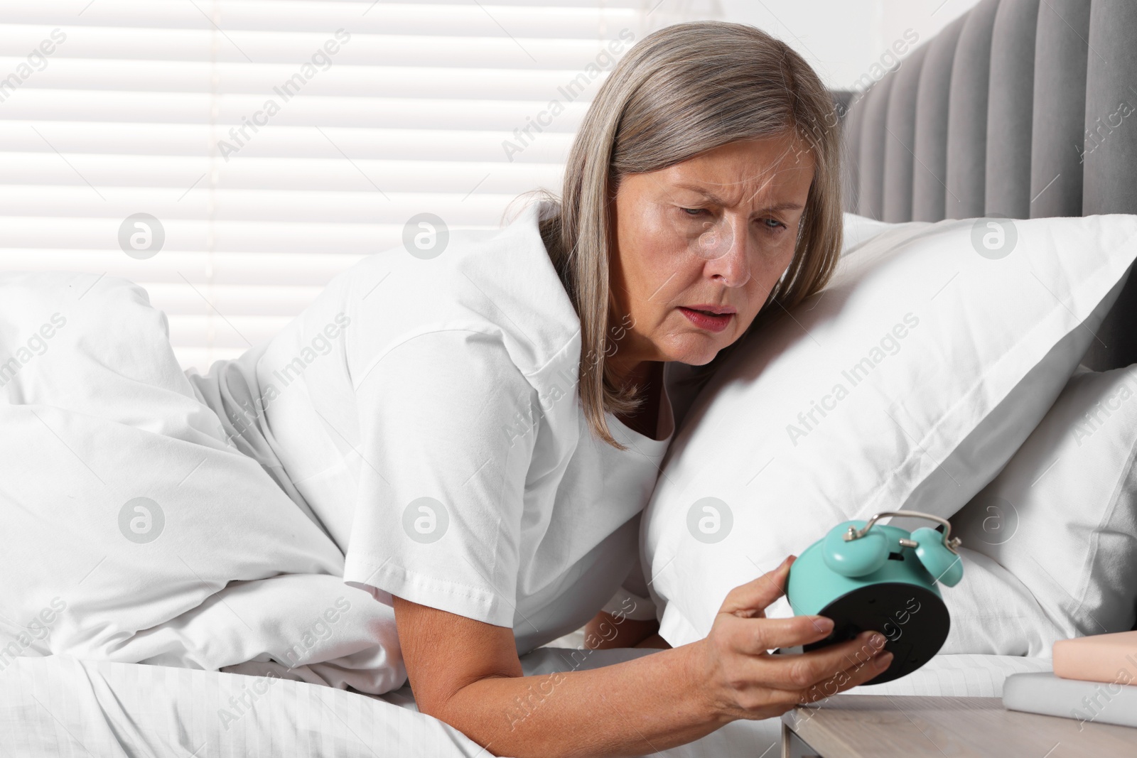 Photo of Overslept woman looking at alarm clock in bed indoors