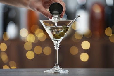 Photo of Woman making tasty martini cocktail with olives at wooden table on blurred background, closeup. Bokeh effect