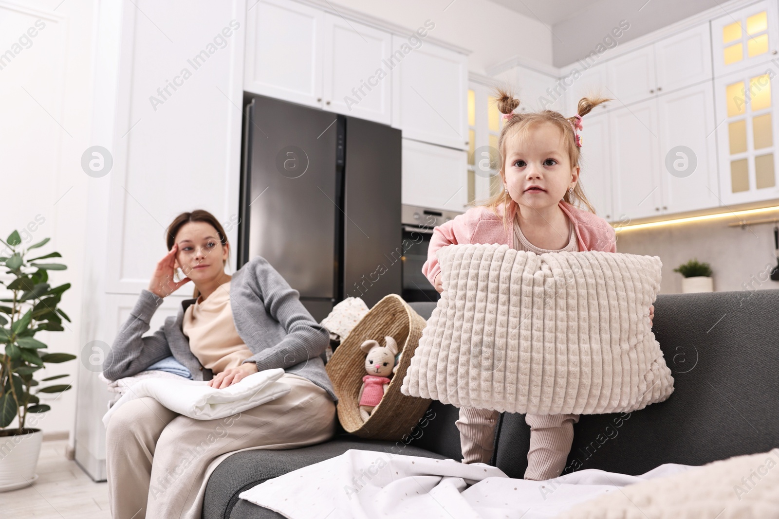 Photo of Tired housewife with her little daughter among messy laundry on sofa at home