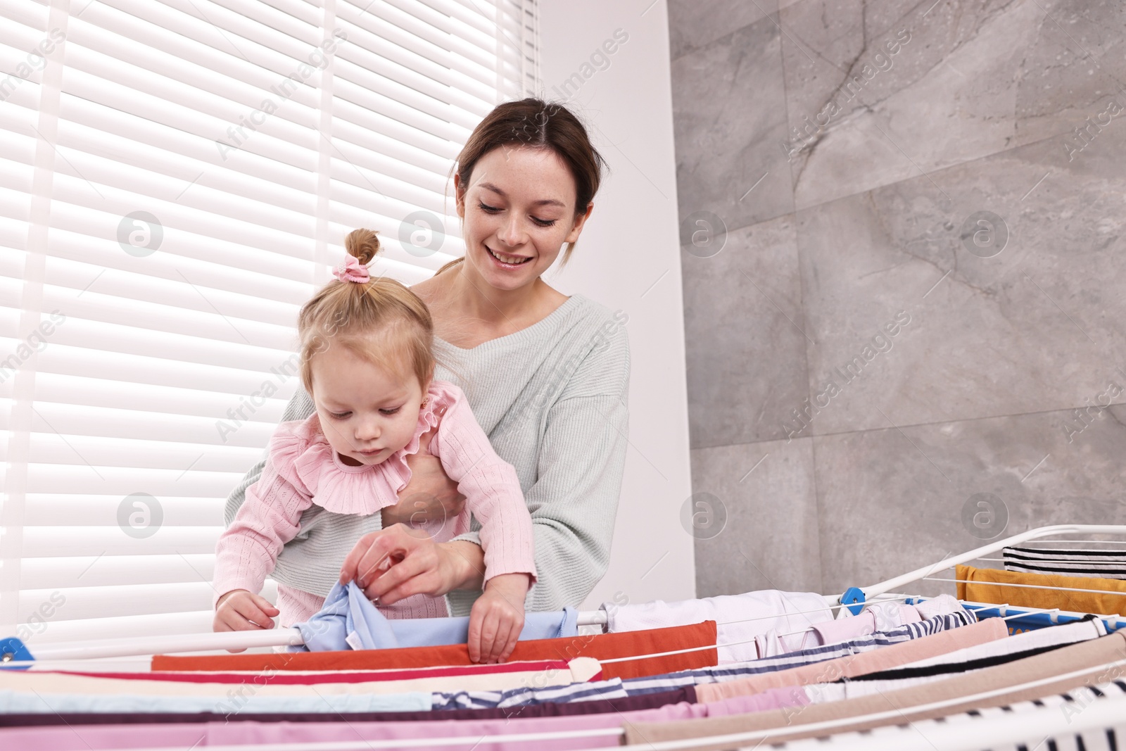 Photo of Smiling housewife with her daughter hanging clothes on drying rack at home