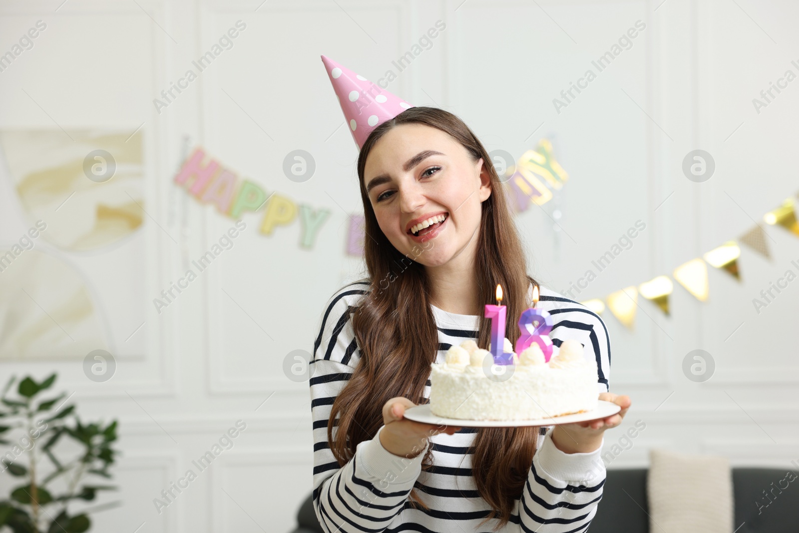 Photo of Coming of age party - 18th birthday. Happy young woman in hat holding tasty cake with number shaped candles at home