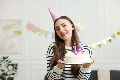 Photo of Coming of age party - 18th birthday. Happy young woman in hat holding tasty cake with number shaped candles at home