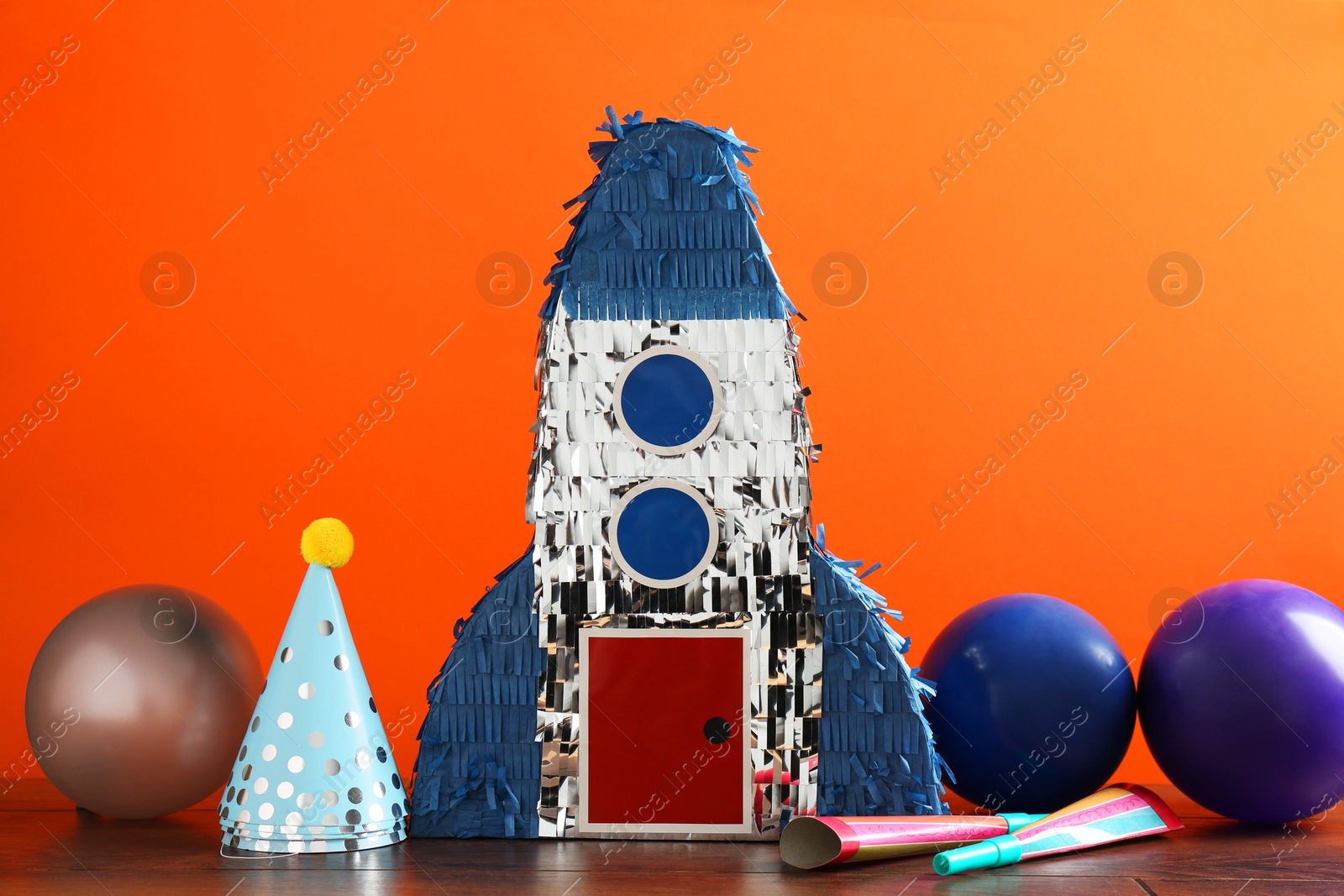 Photo of Bright pinata in shape of spaceship and party accessories on table against orange background