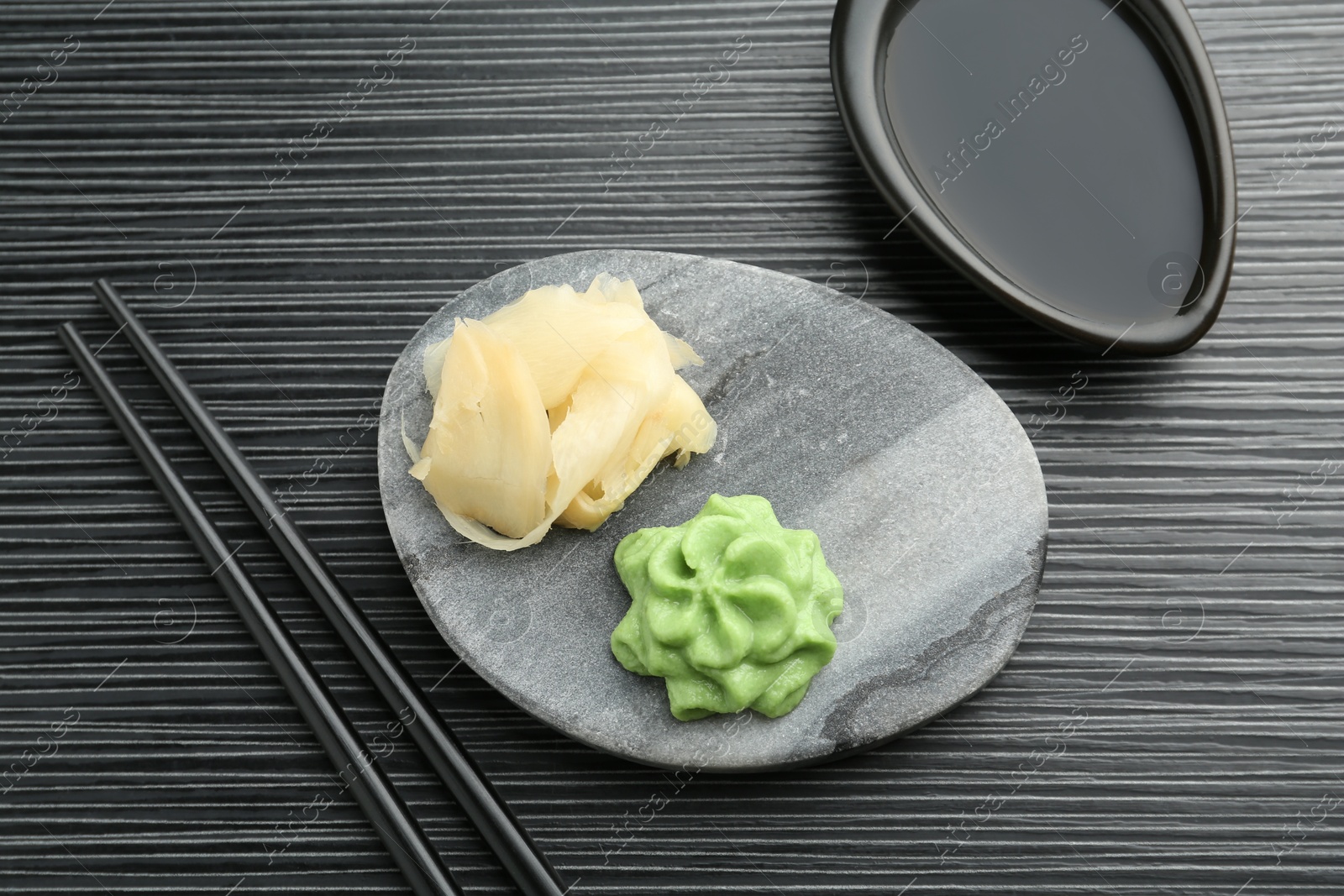 Photo of Hot wasabi paste, soy sauce, ginger and chopsticks on black textured table, flat lay