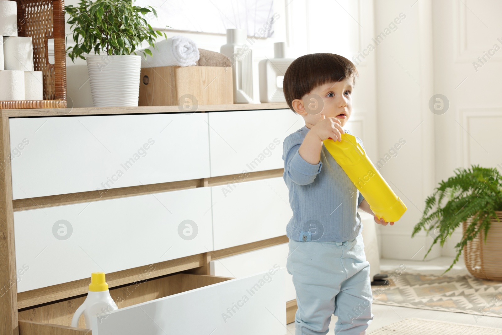 Photo of Little boy with bottle of detergent near cabinet at home. Child in danger