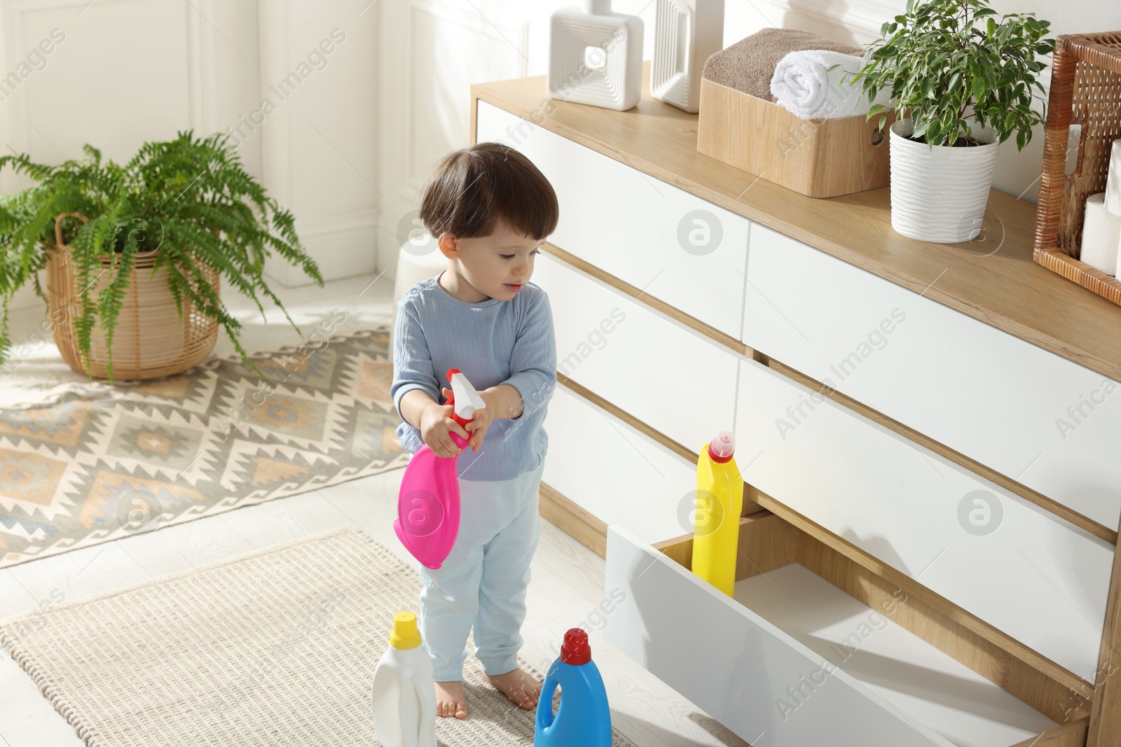 Photo of Little boy playing with bottle of detergent near cabinet at home. Child in danger