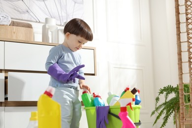 Photo of Little boy playing with glove and bottles of detergents near cabinet at home. Child in danger