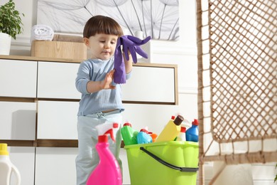 Photo of Little boy playing with glove and bottles of detergents near cabinet at home. Child in danger