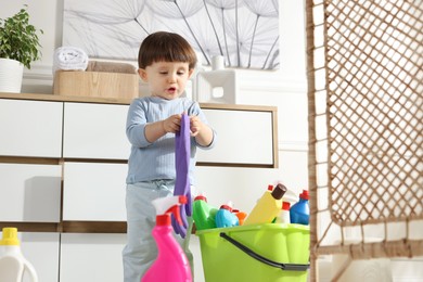 Photo of Little boy playing with glove and bottles of detergents near cabinet at home. Child in danger