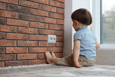 Photo of Little boy sitting near electrical socket at home, space for text. Child in danger