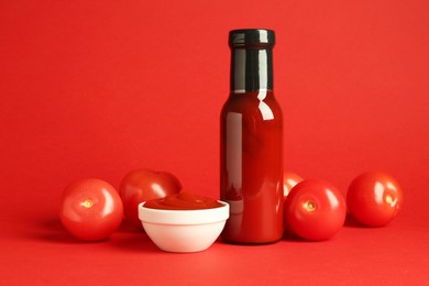 Photo of Ketchup in glass bottle, bowl and fresh tomatoes on red background