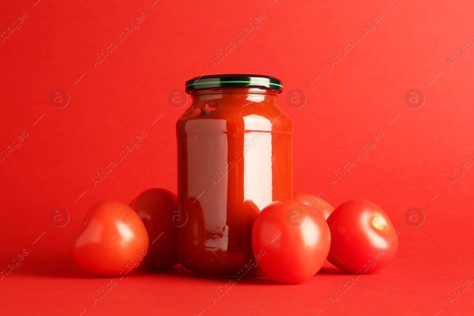 Photo of Ketchup in glass jar and fresh tomatoes on red background