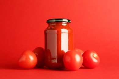 Photo of Ketchup in glass jar and fresh tomatoes on red background