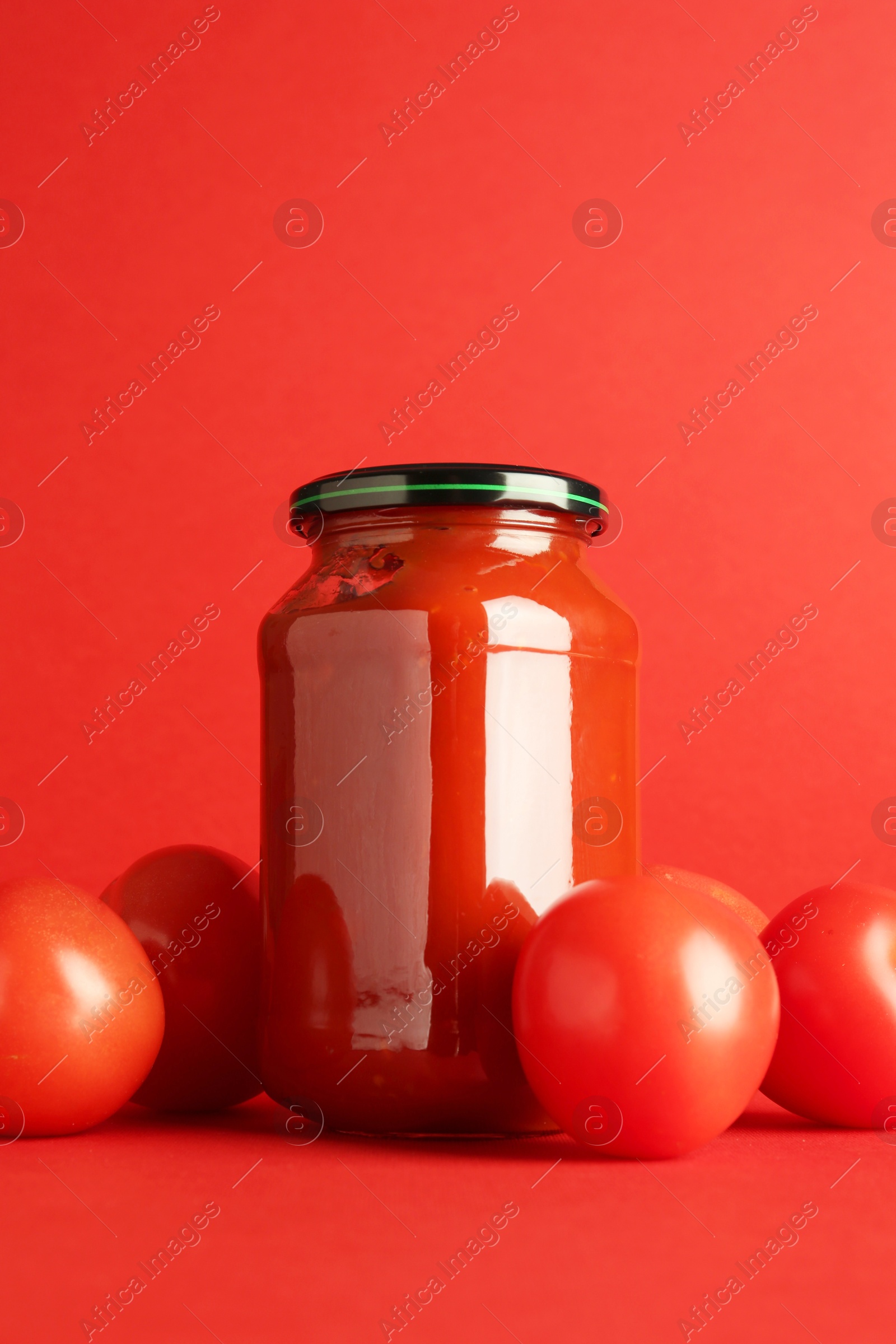 Photo of Ketchup in glass jar and fresh tomatoes on red background