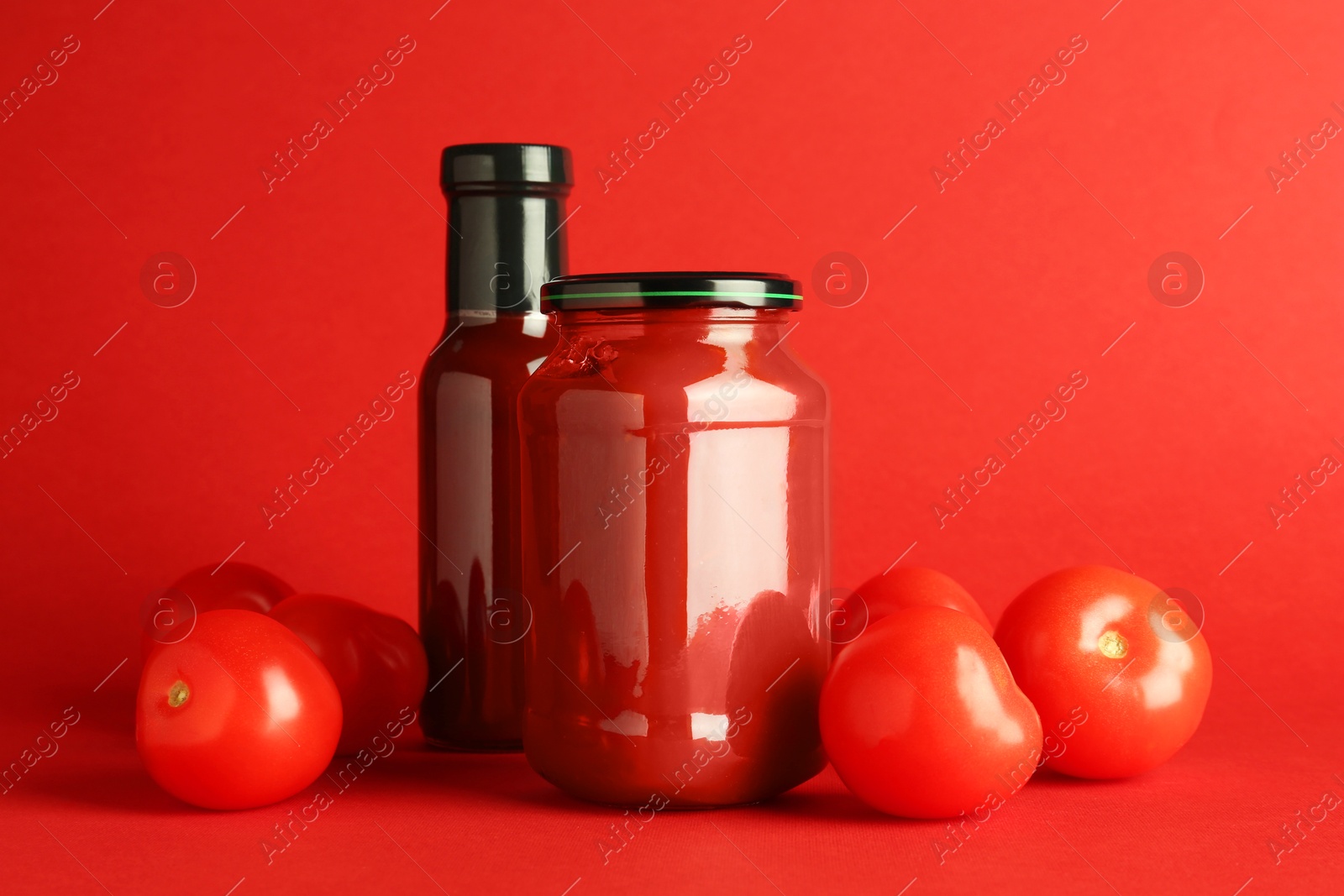 Photo of Ketchup in glass bottle, jar and fresh tomatoes on red background