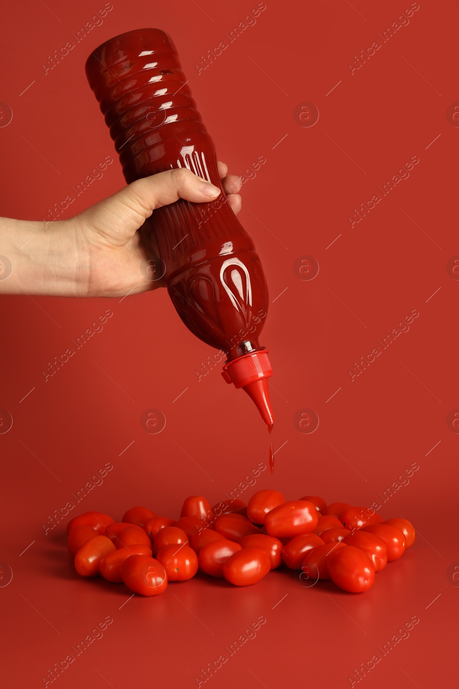 Photo of Woman squeezing ketchup from bottle over tomatoes on red background, closeup