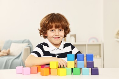 Photo of Little boy stacking colorful cubes at white table indoors