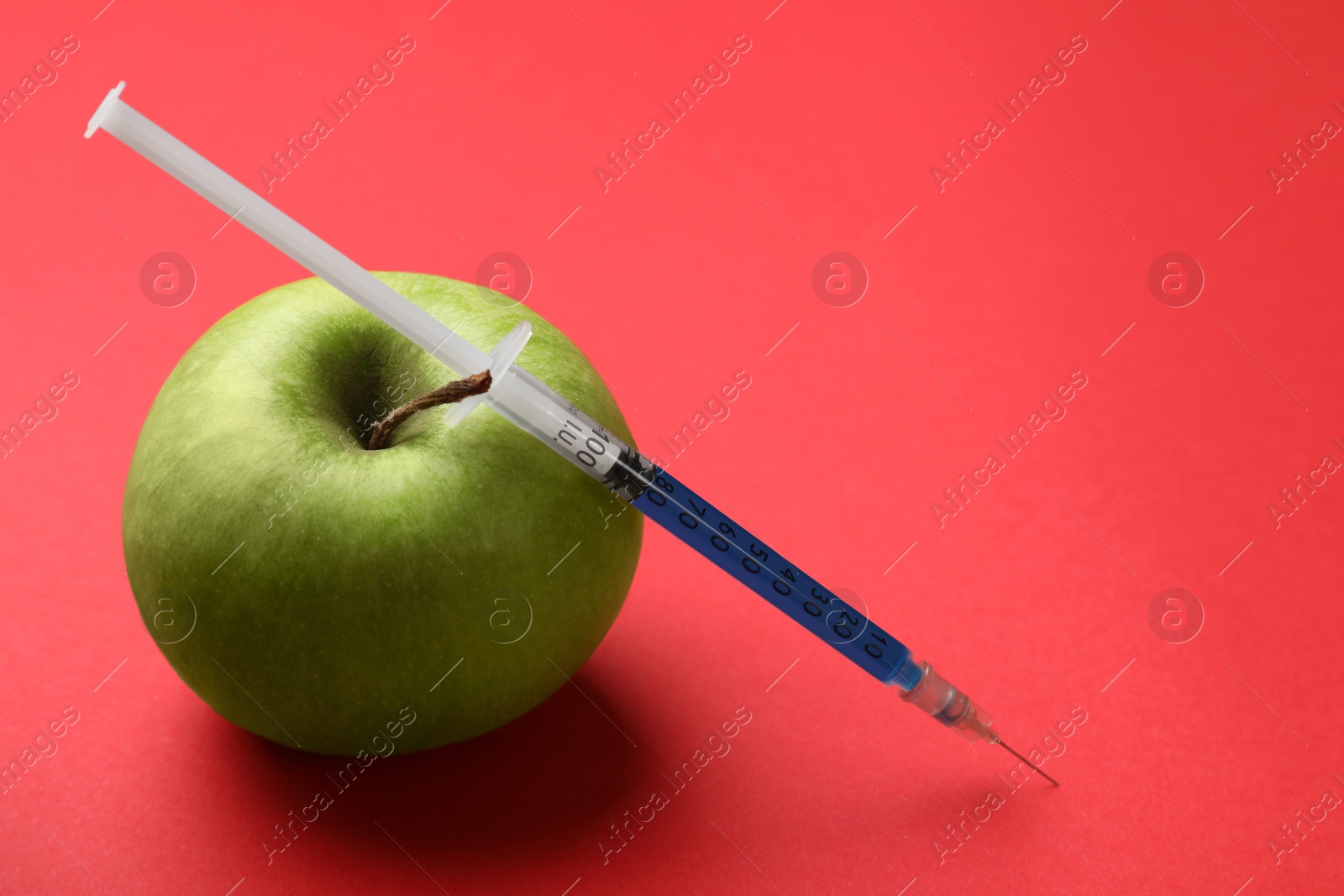 Photo of GMO concept. Green apple and syringe with liquid on red background, closeup