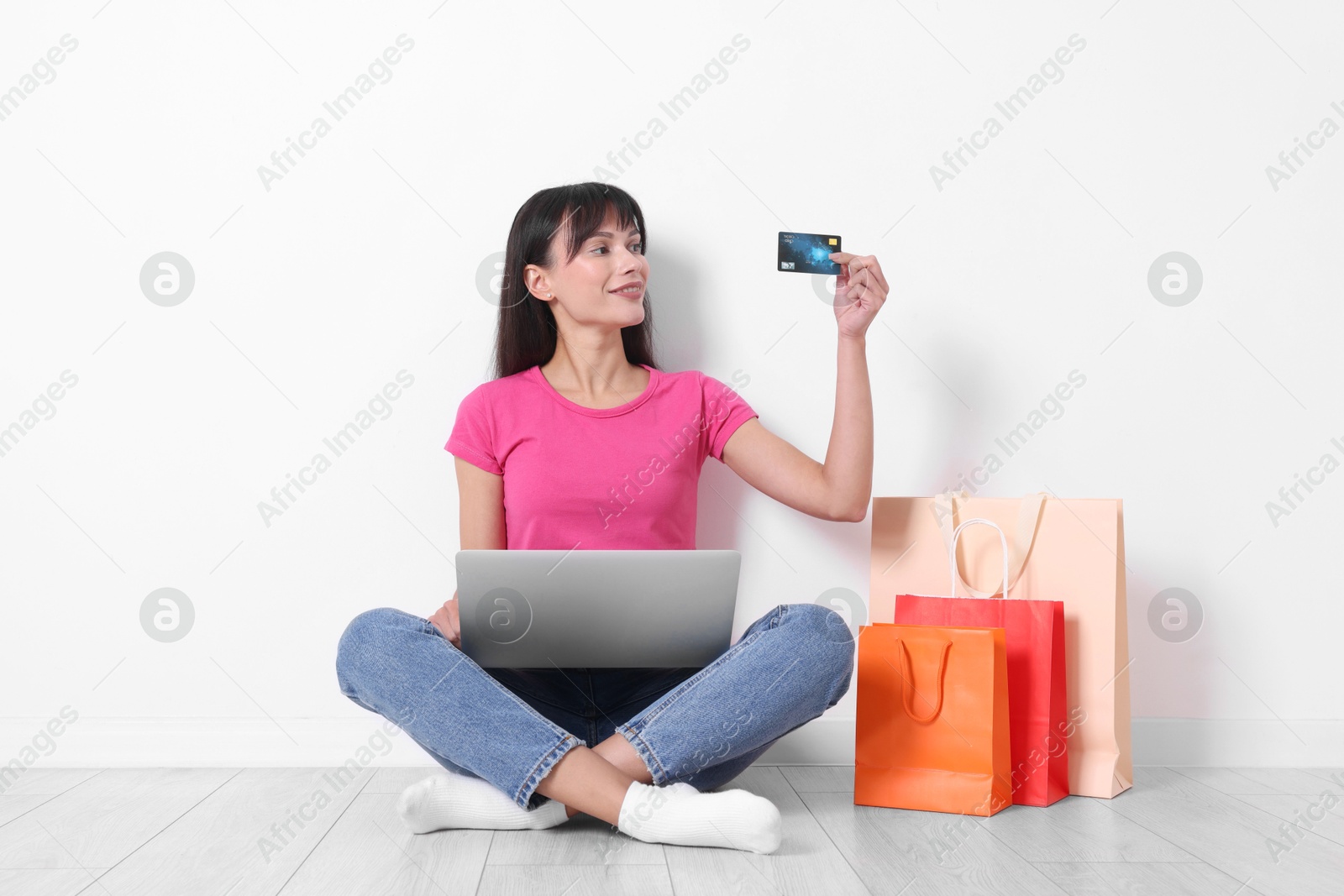 Photo of Internet shopping. Happy woman with credit card, laptop and colorful bags sitting on floor near white wall