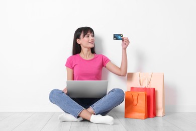 Photo of Internet shopping. Happy woman with credit card, laptop and colorful bags sitting on floor near white wall