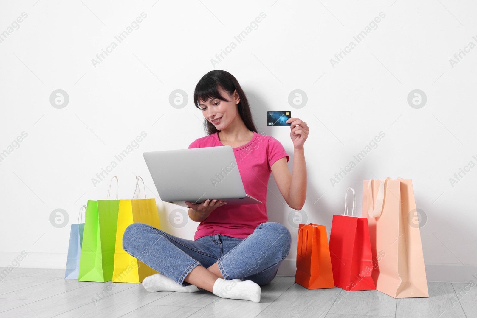 Photo of Internet shopping. Happy woman with credit card, laptop and colorful bags sitting on floor near white wall