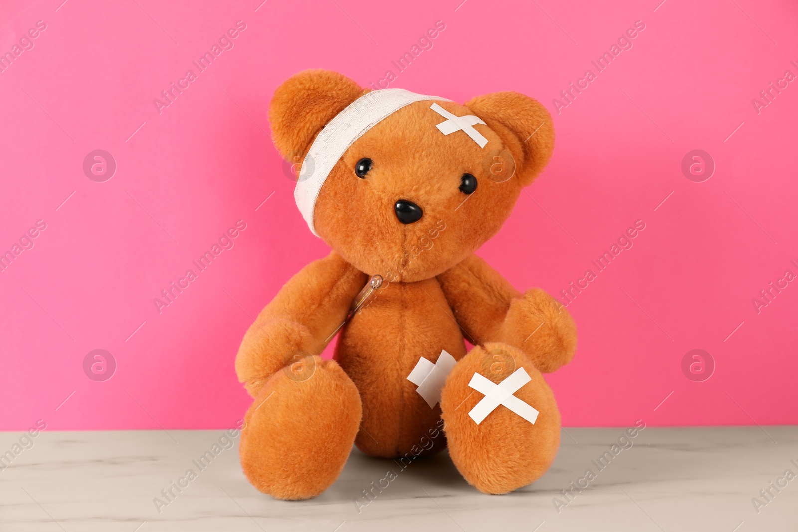 Photo of Teddy bear with bandage, sticking plasters and thermometer on white marble table against pink background