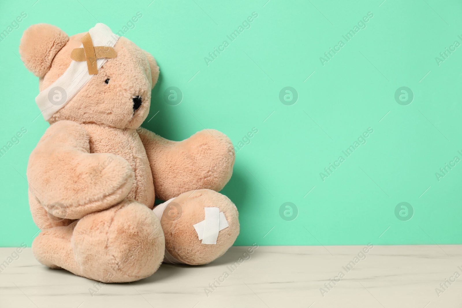 Teddy bear with bandages and sticking plasters on white marble table against turquoise background, space for text Photo of Teddy bear with bandages and sticking plasters on white marble table against turquoise background, space for text