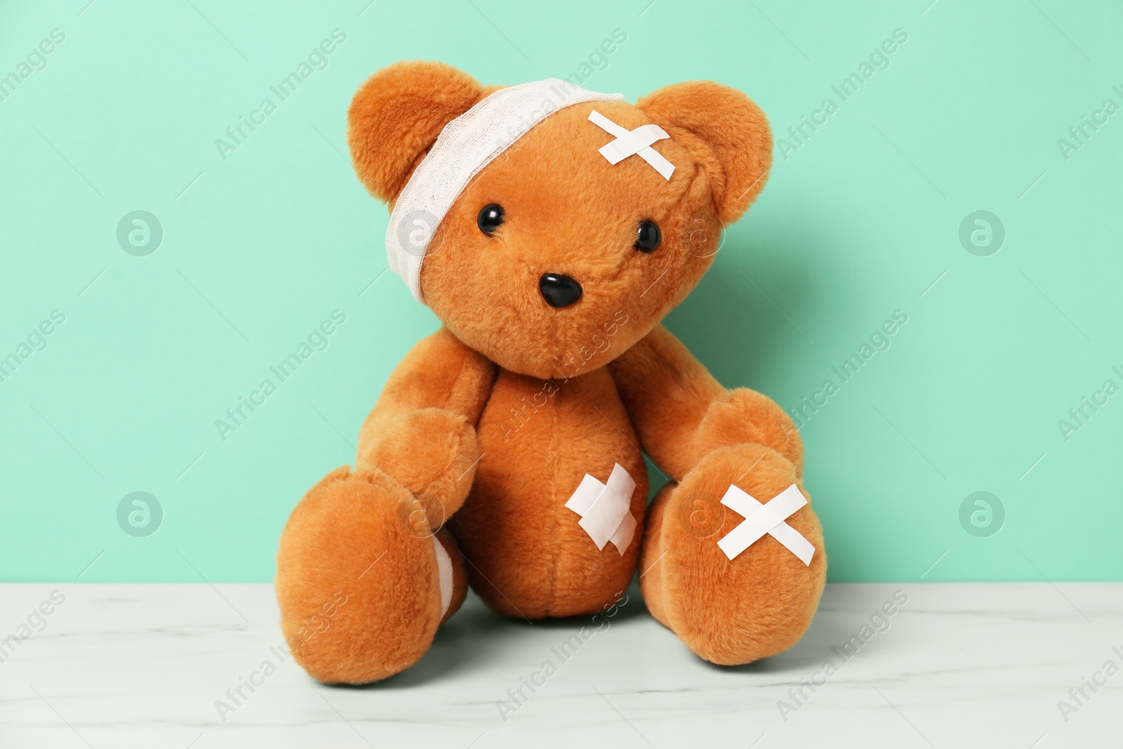 Teddy bear with bandages and sticking plasters on white marble table against turquoise background Photo of Teddy bear with bandages and sticking plasters on white marble table against turquoise background