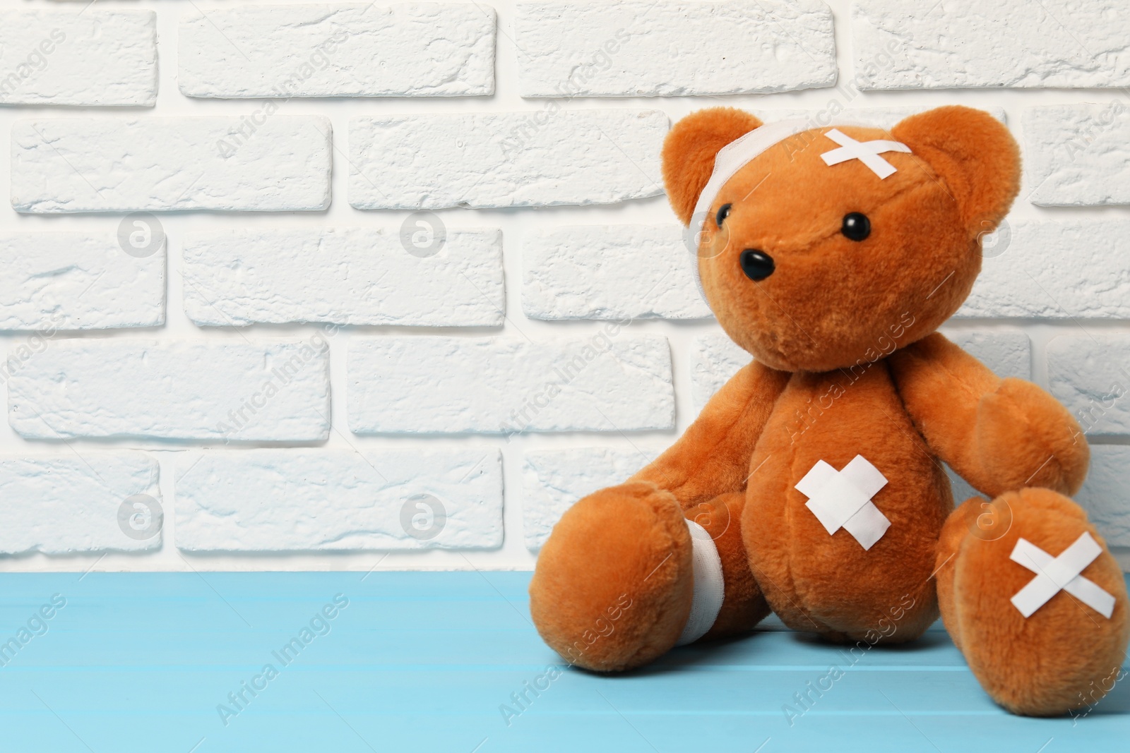 Teddy bear with bandages and sticking plasters on light blue wooden table against white brick wall, space for text Photo of Teddy bear with bandages and sticking plasters on light blue wooden table against white brick wall, space for text